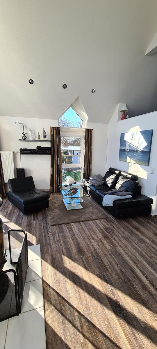 Wohnzimmer mit schwarzen Ledermöbeln, Glas-Tisch und Holzboden. Fenster mit Vorhängen.