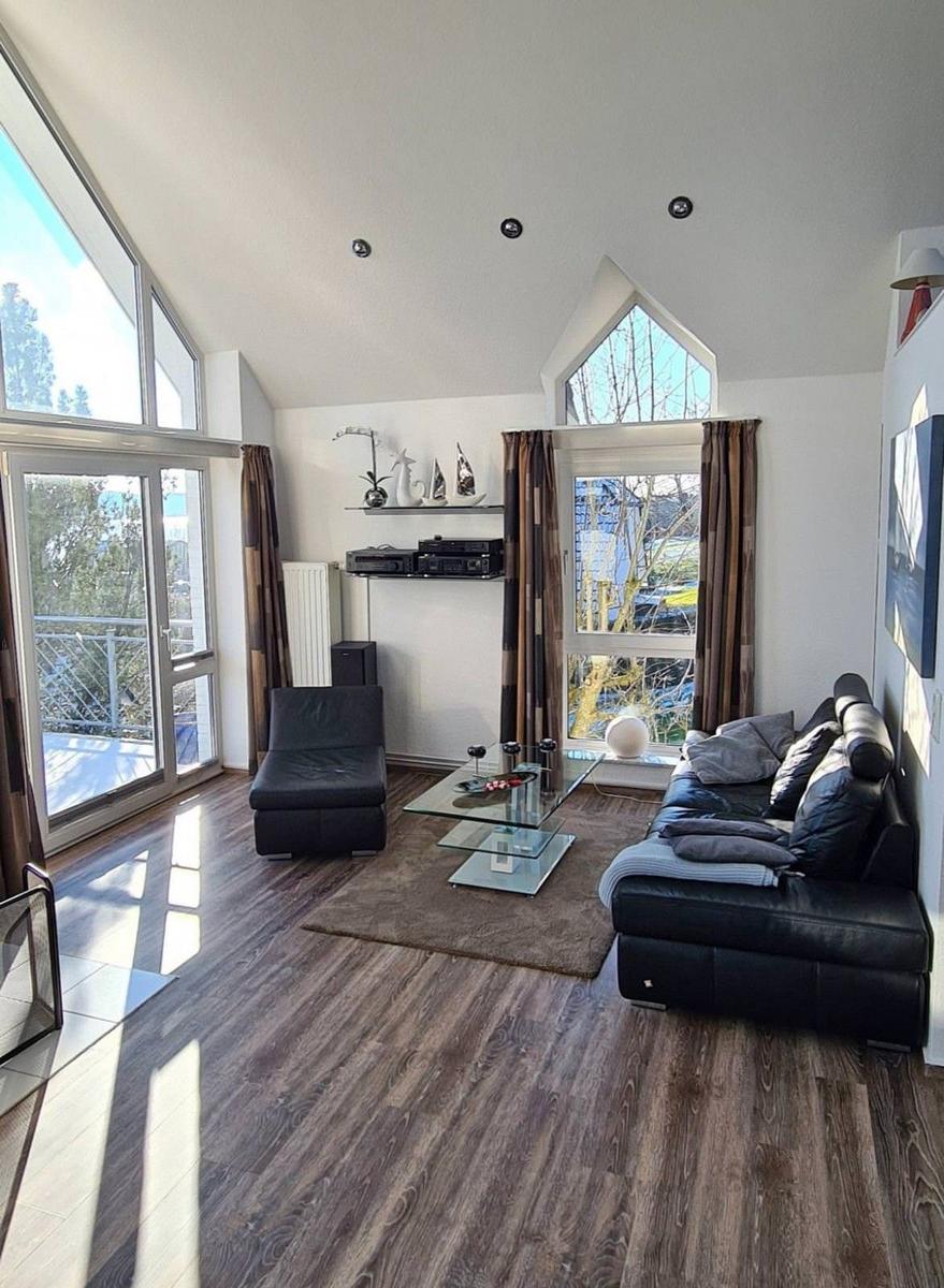 Wohnzimmer mit schwarzem Sofa, Glas-Tisch und großen Fenstern.