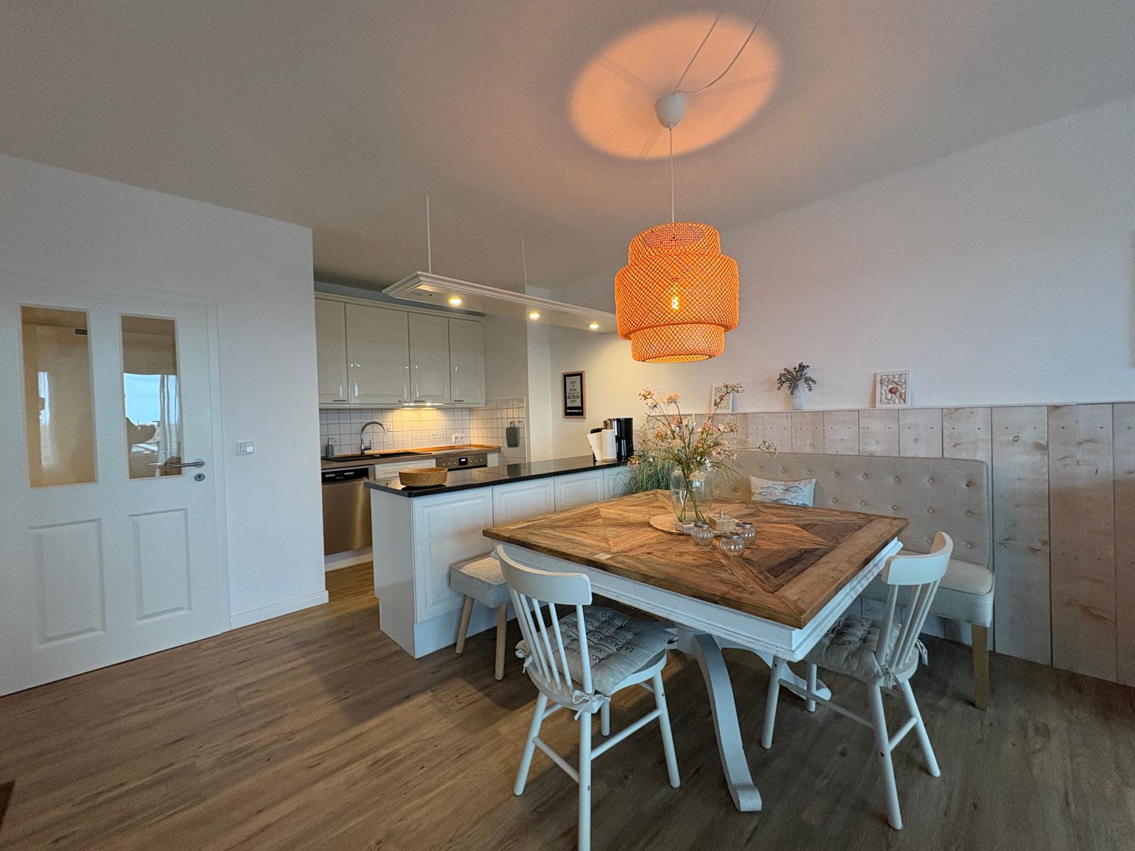 Essbereich mit Holztisch, weißen Stühlen und orangefarbener Hängelampe. Küche im Hintergrund.