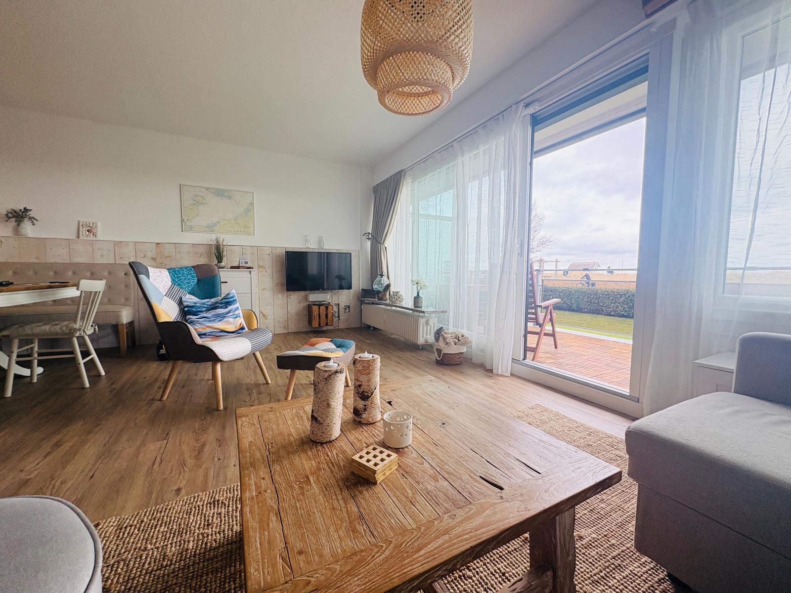 Wohnzimmer mit Holzboden, Sitzgelegenheiten und großem Fenster zum Balkon.