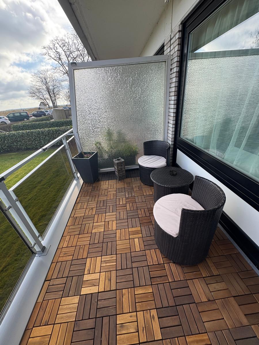 Terrasse mit zwei Stühlen und Tisch, Holzbelag, Glasgeländer und Sicht auf Grünfläche.