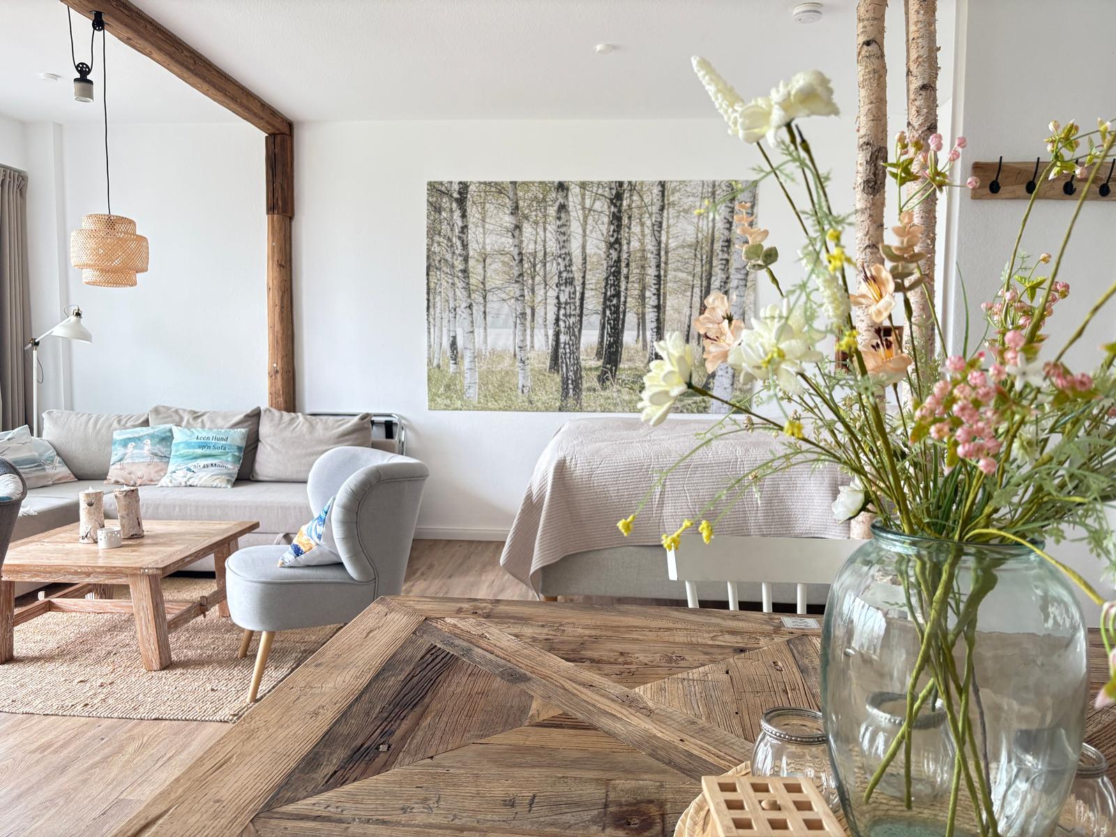 Wohnraum mit Sofa, Stuhl und Holztisch. Wandbild zeigt Birkenwald. Blumen im Glasvasen.