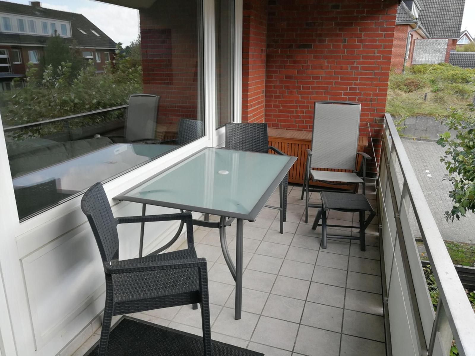 Terrasse mit Glas-Tisch und Stühlen, roter Backsteinwand, Blick auf Grünfläche und Straße.