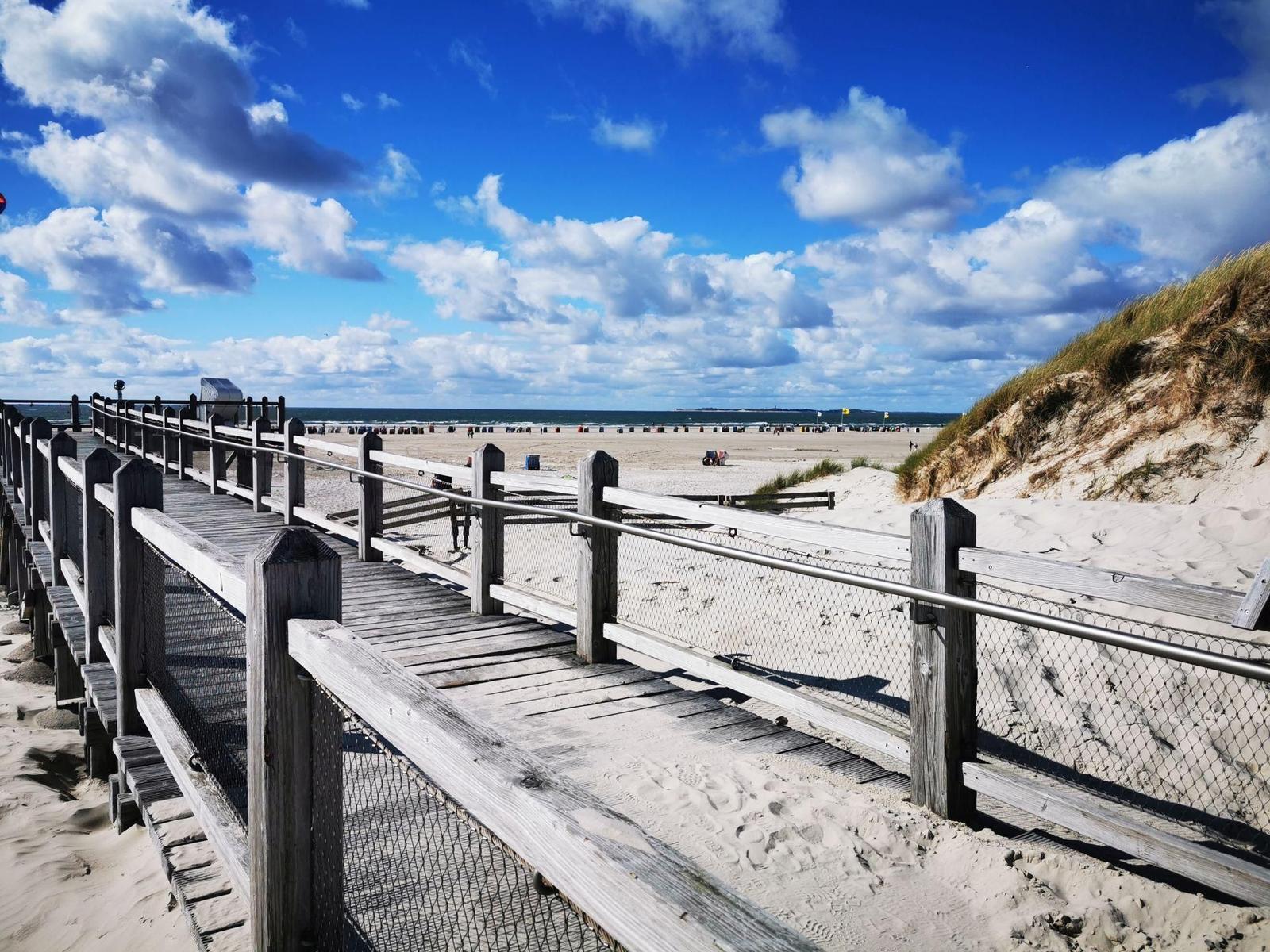 Holzbrücke führt zum Strand. Himmel blau mit weißen Wolken. Dünen und Sandstrand im Hintergrund.