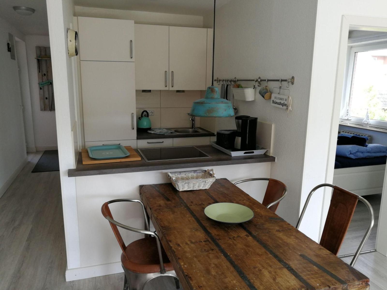 Küche mit Esstisch und Stühlen. Im Hintergrund ist ein Schlafzimmer mit Fenster sichtbar.