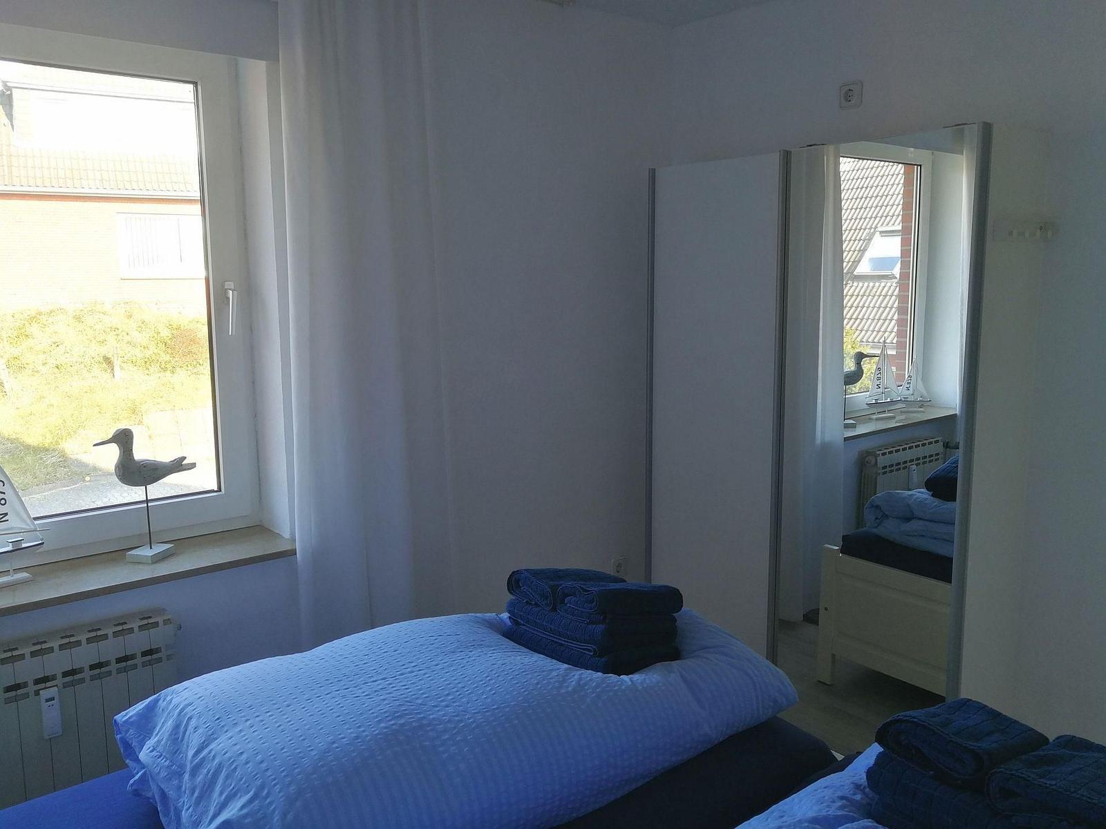Zimmer mit Bett, Spiegel und Fenster. Auf dem Fensterbrett stehen dekorative Gegenstände.