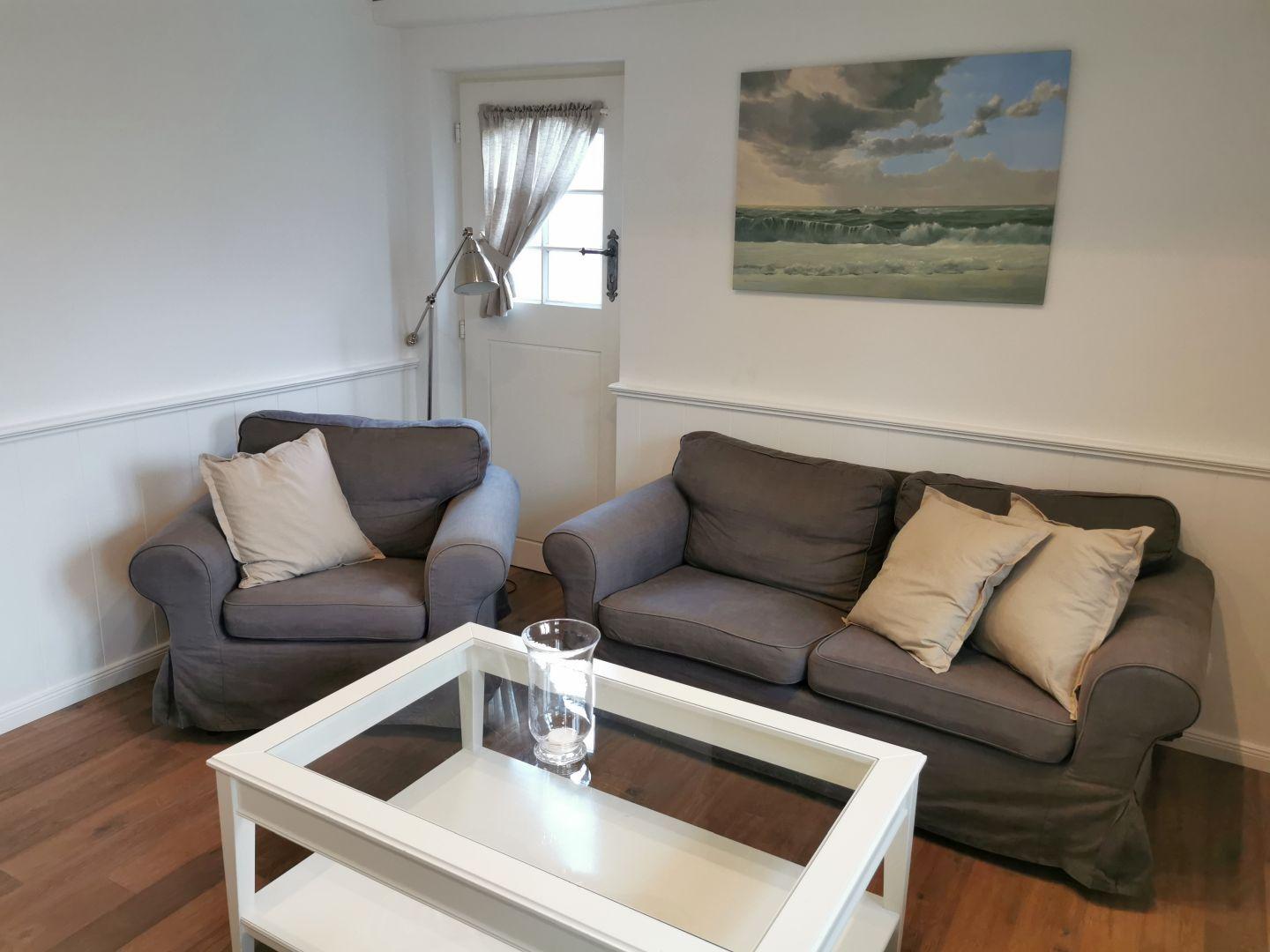 Wohnzimmer mit grauem Sofa, Sessel und Glas-Tisch. Wandbild zeigt Meereslandschaft.