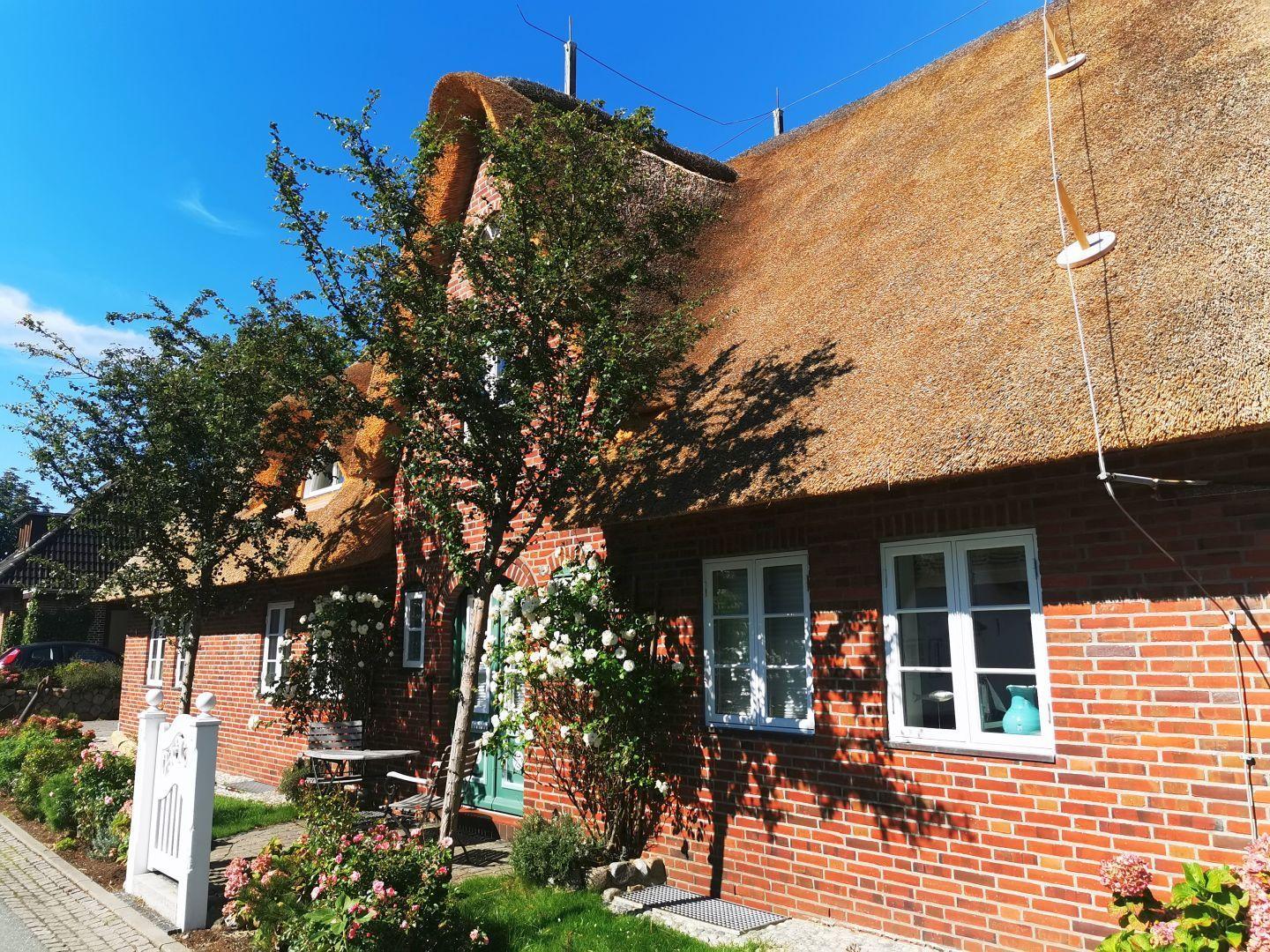 Rotes Backsteinhaus mit strohgedecktem Dach und weißen Fenstern. Garten mit Blumen und Tisch.