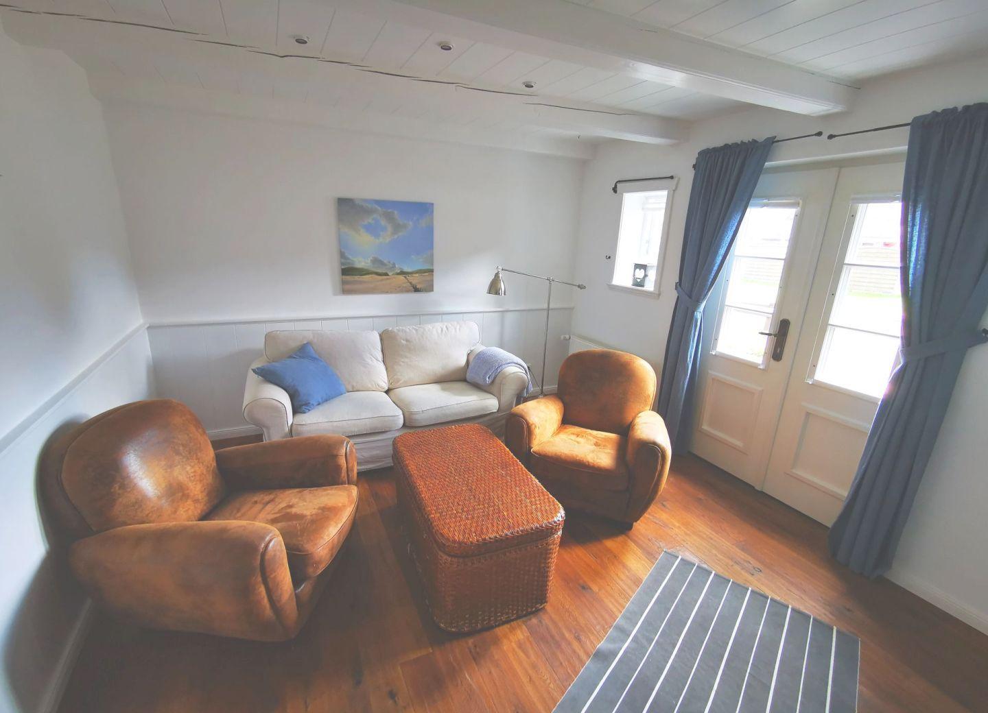 Wohnzimmer mit Sofa, zwei Ledersessel und Holzboden. Fenster mit blauen Vorhängen.