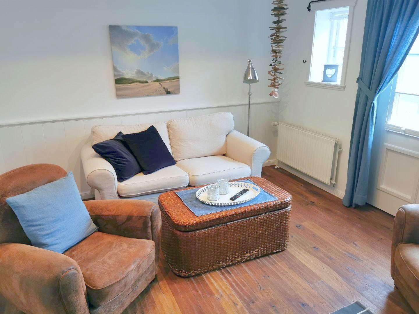 Wohnzimmer mit Sofa, Sessel und Kaffeetisch. Holzboden und Fenster mit Vorhängen.