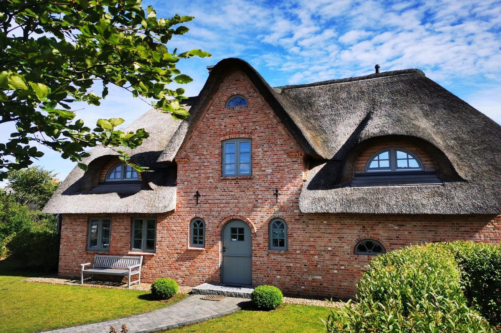 Rotes Backsteinhaus mit Strohdach und grüner Tür. Garten mit Bank und Beerensträuchern.