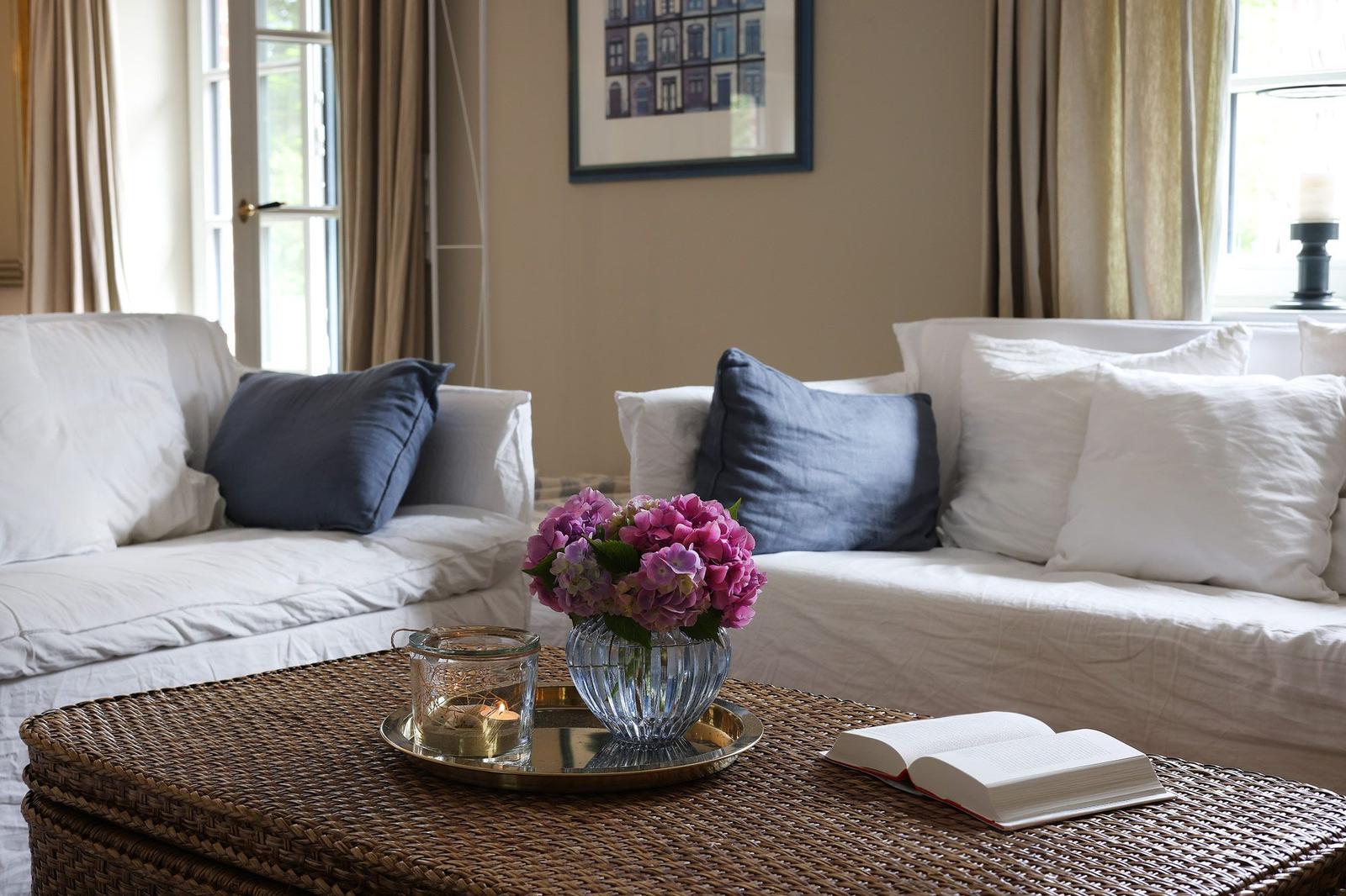 Wohnzimmer mit weißen Sofas, blauen Kissen und einem Korbtablett mit Blumen und Buch.