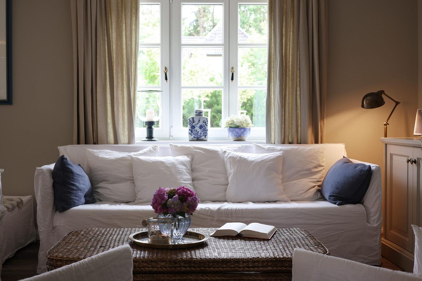 Ein Wohnzimmer mit Sofa, Tisch und Fenster. Auf dem Tisch steht ein Buch und Blumen.