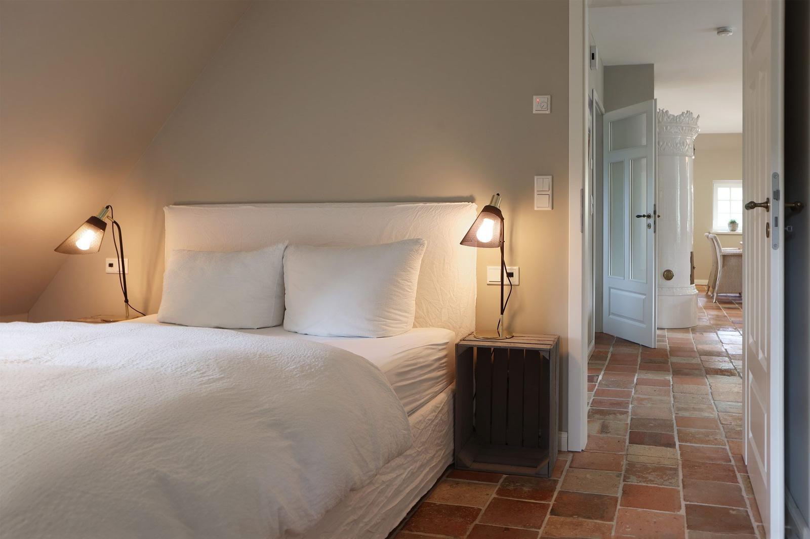 Schlafzimmer mit Bett, Nachttisch und Stehlampe. Blick in den Flur mit Terrakottaboden.