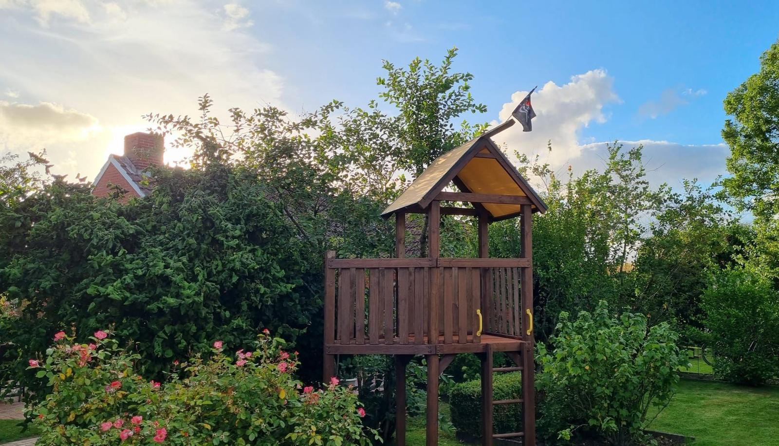 Holzspielhaus mit Flagge im Garten, umgeben von Bäumen und Rosen.