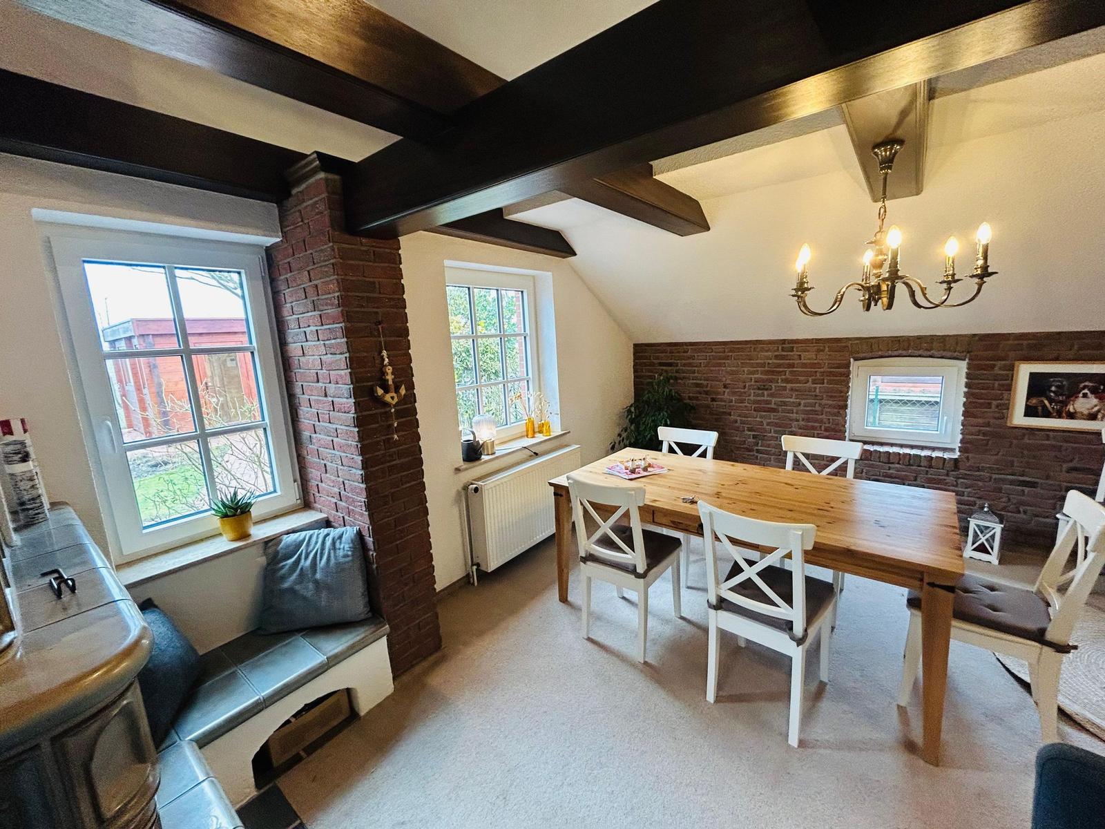 Esszimmer mit Holztisch, weißen Stühlen und Backsteinwand. Deckenbalken und Kronleuchter sichtbar.