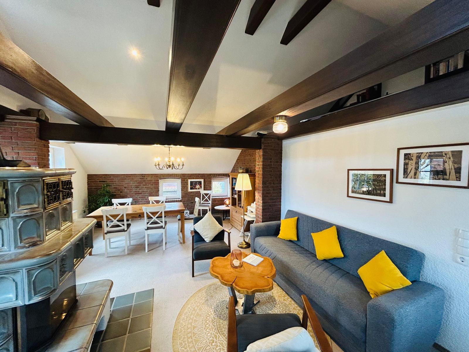 Wohn- und Essbereich mit Holzbalken, Sofa mit gelben Kissen und Kachelofen.