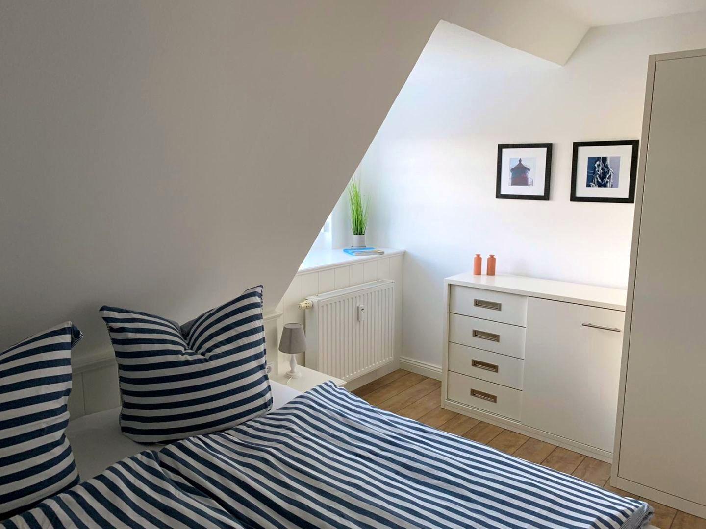 Schlafzimmer mit Dachschräge, Bett mit Streifen, Schrank und Bilder an der Wand.