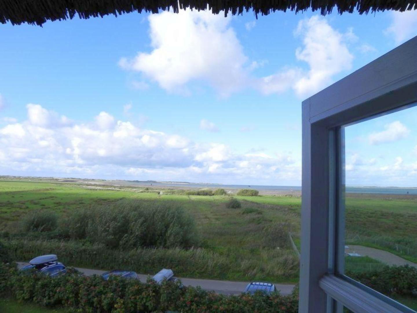 Blick aus dem Fenster auf grünes Feld und Meer unter blauem Himmel mit weißen Wolken.