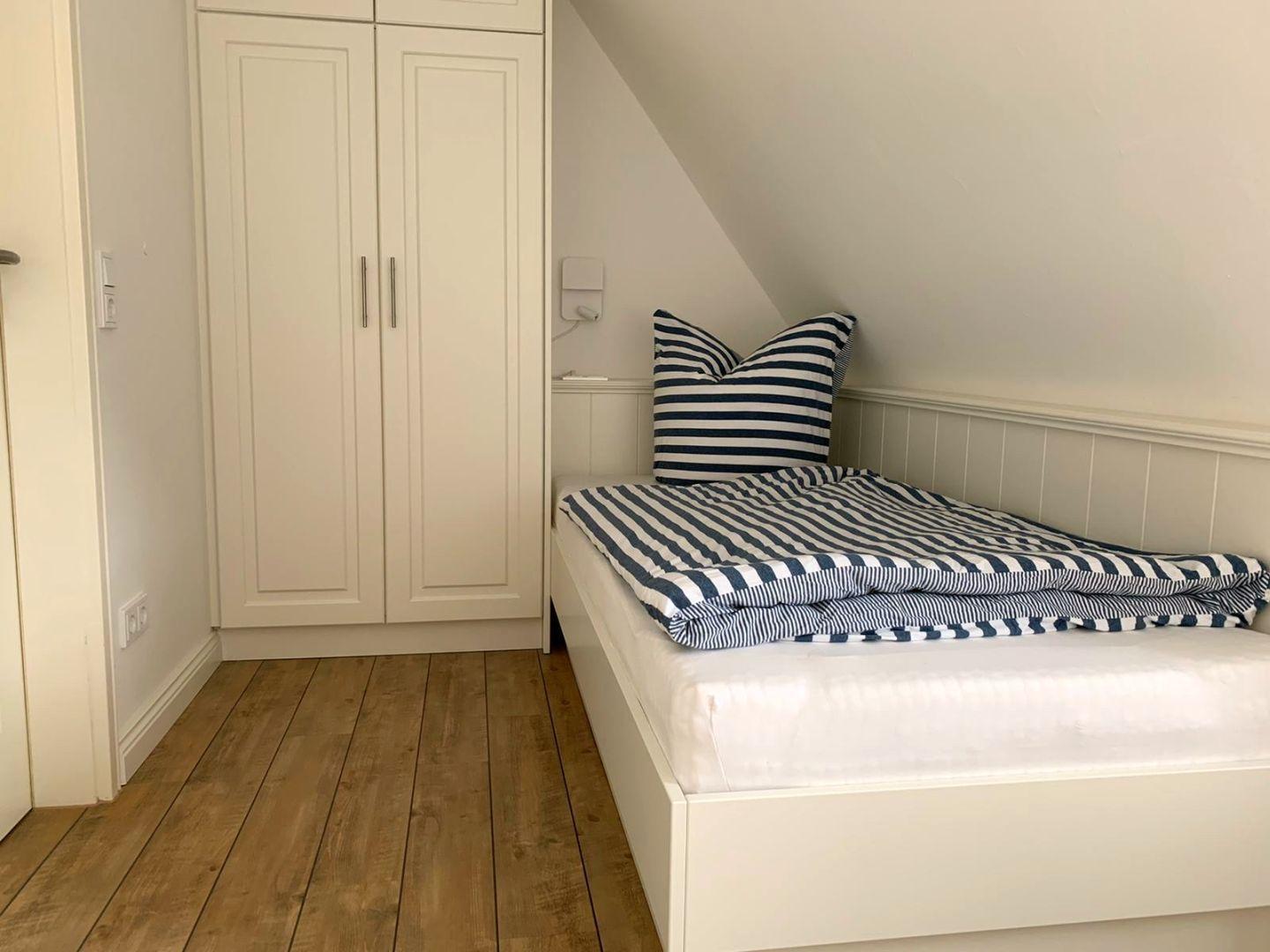 Schlafzimmer mit weißem Bett und Streifenbezug, Holzboden und weißen Schränken unter schräger Decke.