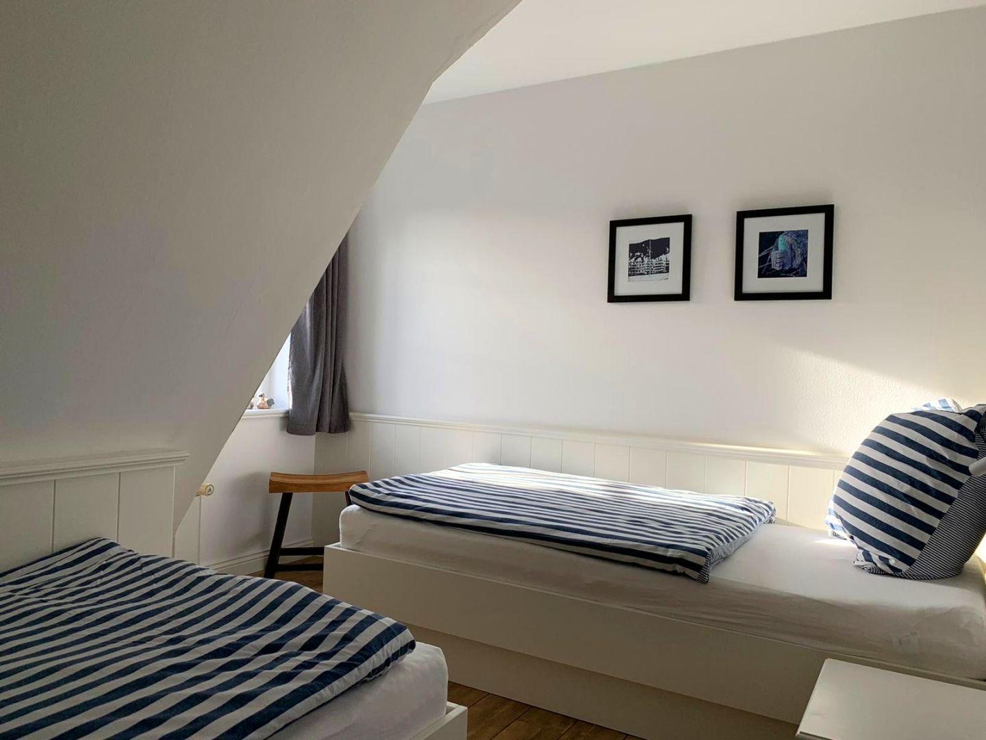 Zimmer mit zwei Betten, schräger Decke und Fenster mit Vorhang.