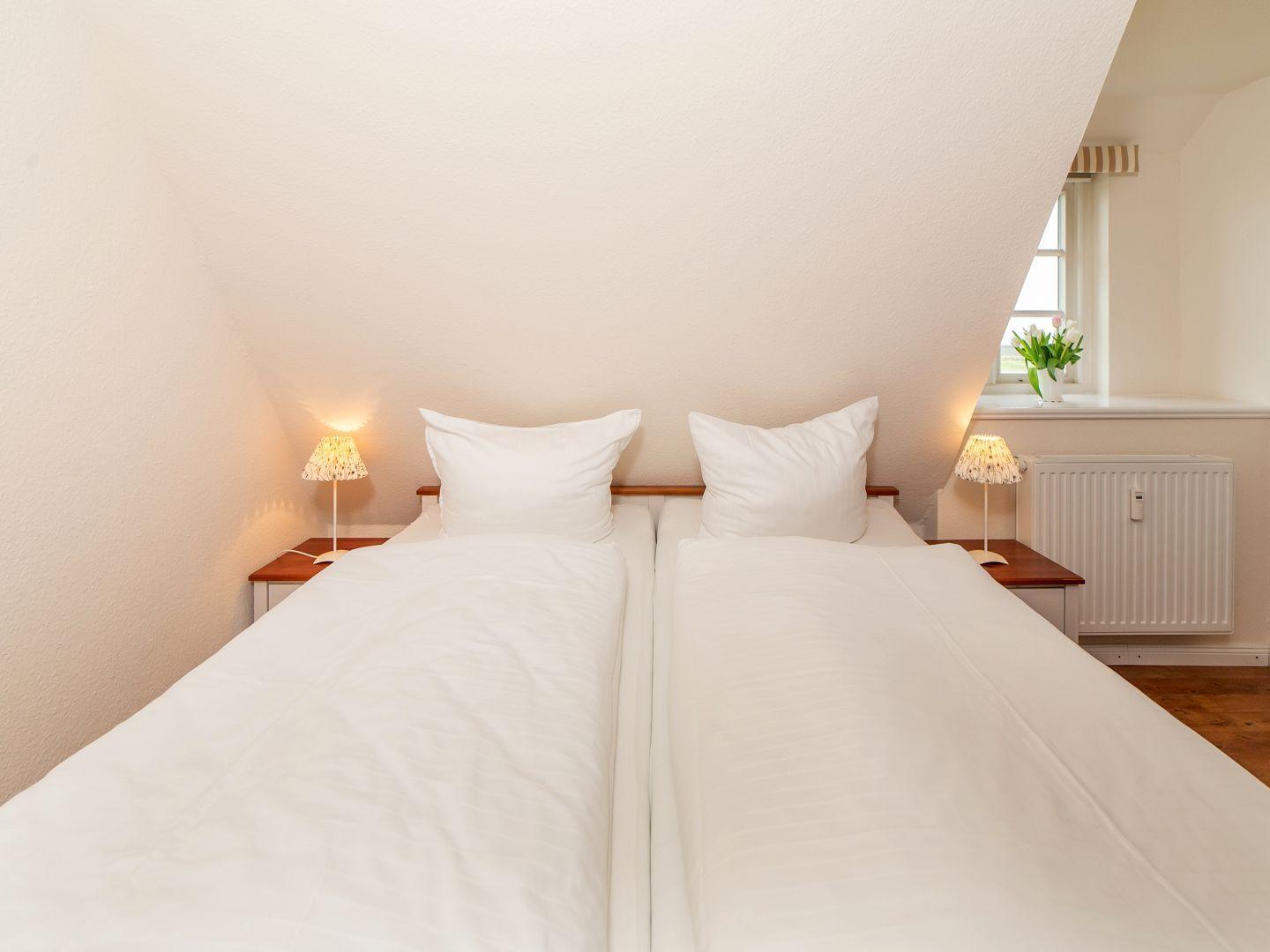 Doppelbett mit weißen Laken, zwei Nachttische mit Lampen und Fenster mit Blumen.