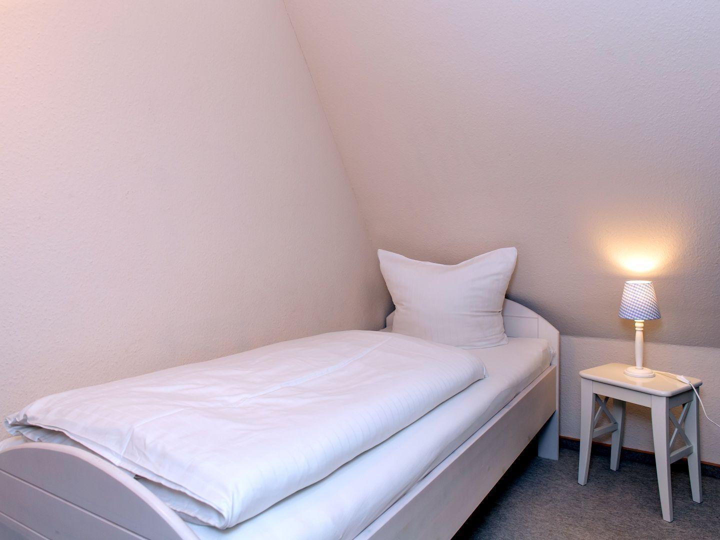 Ein Schlafzimmer mit Einzelbett, Nachttisch und Lampe. Die Wände sind hell gestrichen.
