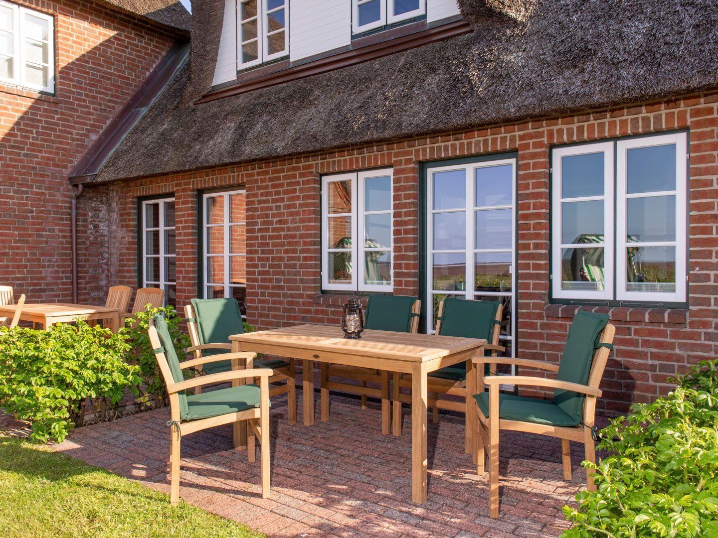 Terrasse mit Holztisch und grünen Stühlen vor einem Backsteinhaus mit Strohdach.