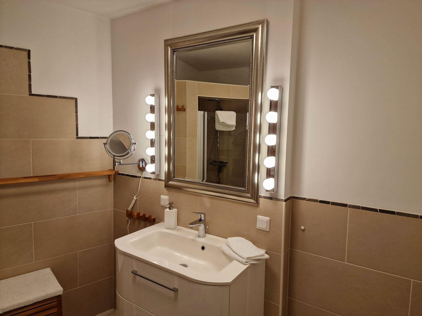 Bad mit Waschbecken, Spiegel mit Licht und Dusche im Hintergrund.