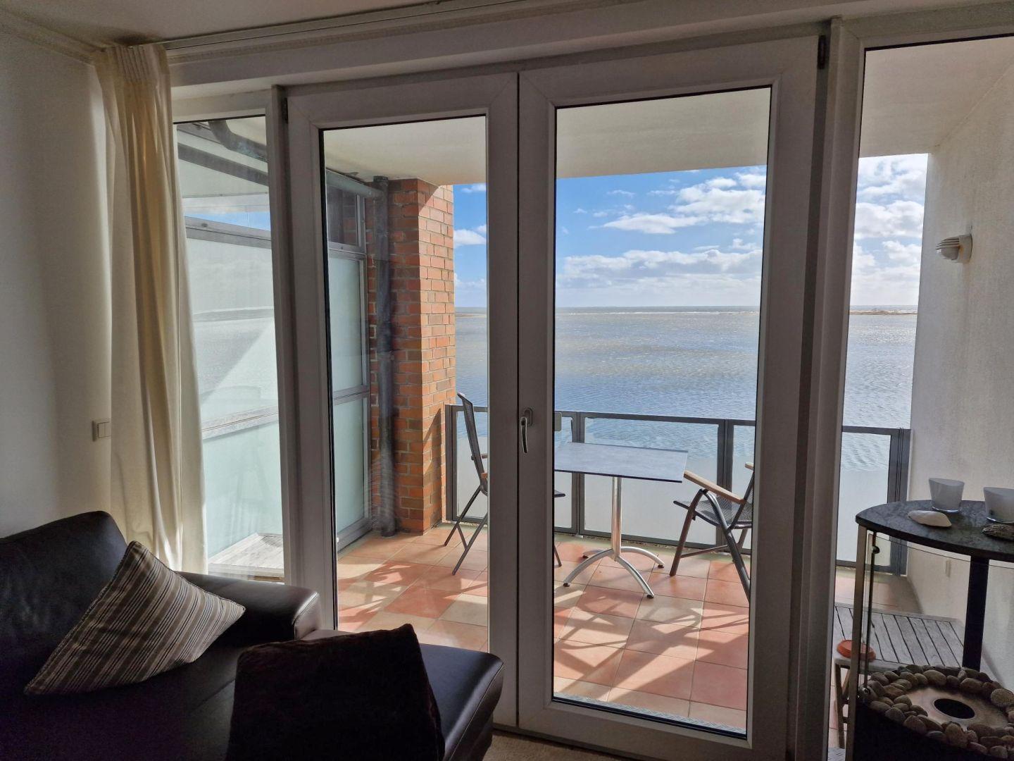 Durch Glastüren führt ein Balkon mit Tisch und Stühlen zum Meer.