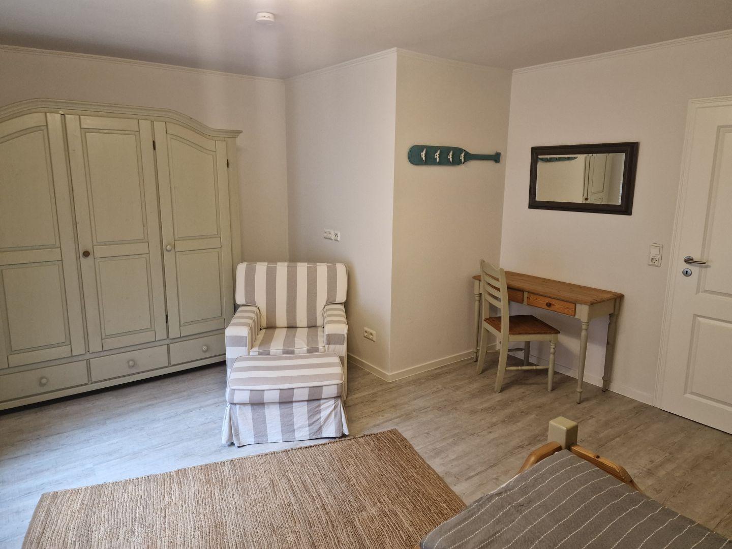 Schlafzimmer mit Kleiderschrank, Sessel, Schreibtisch und Spiegel an der Wand.