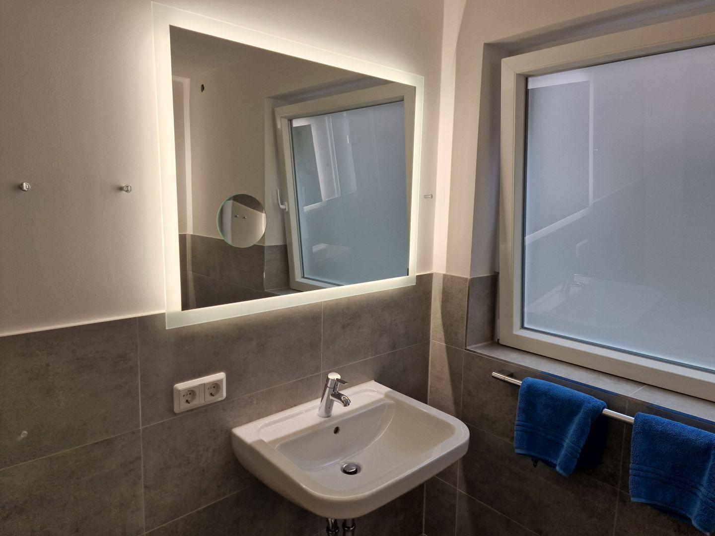 Badezimmer mit Waschbecken, beleuchtetem Spiegel und blauen Handtüchern an der Wand.