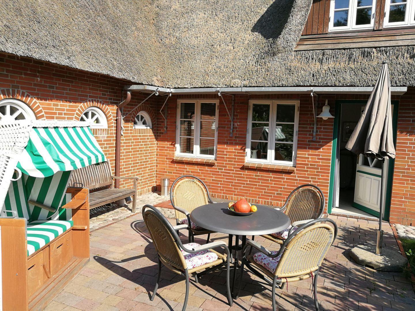 Terrasse mit Tisch, Stühlen und Strandkorb vor einem Haus mit Strohdach.