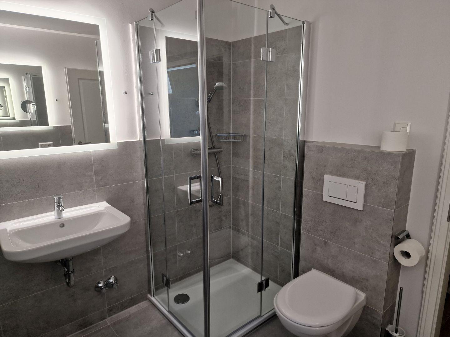 Badezimmer mit Dusche, Waschbecken und Toilette. Fliesen und Spiegel sind modern.