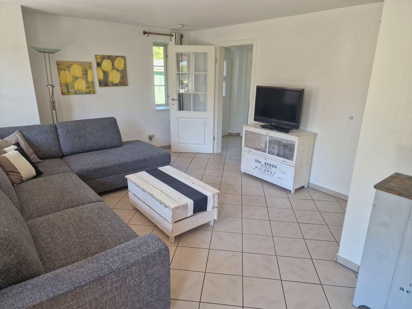 Wohnzimmer mit grauem Sofa, Fernseher und Holztisch auf Fliesenboden.