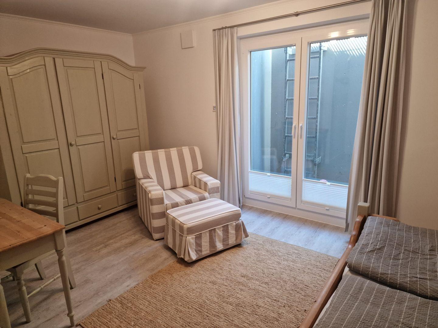 Zimmer mit Schrank, Sessel, Tisch und Fenster zum Balkon.