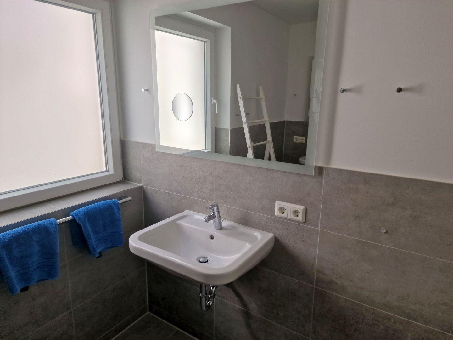 Badezimmer mit Waschbecken, Spiegel und Fenster. Blaue Handtücher hängen an einem Halter.