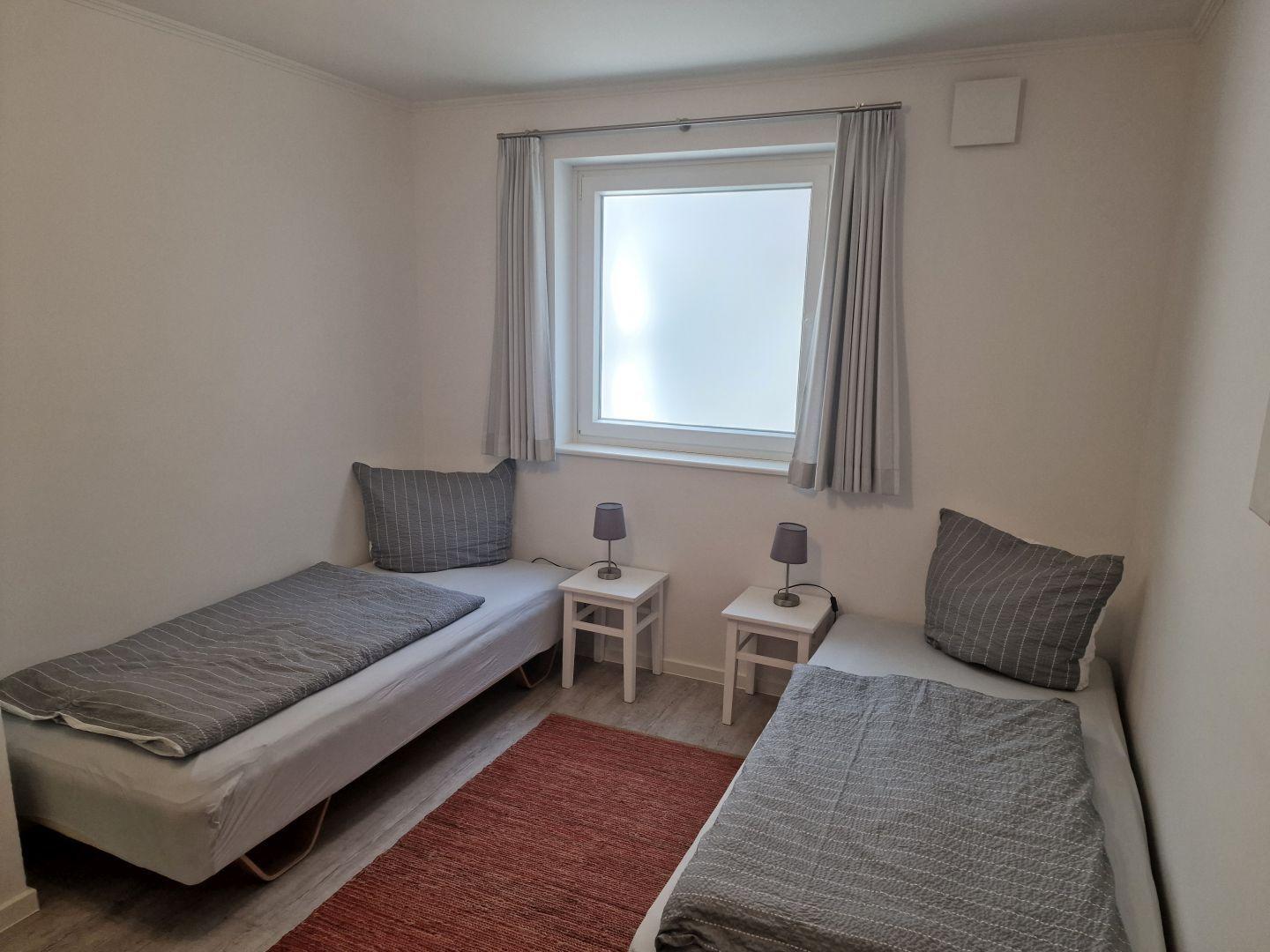 Zwei Einzelbetten mit grauer Bettwäsche, zwei Nachttische und ein Fenster mit Vorhängen.