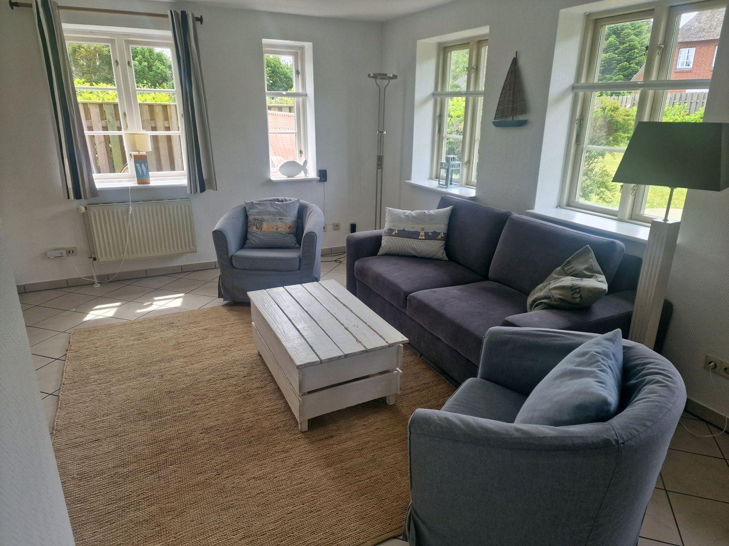Wohnzimmer mit Sofa, zwei Sesseln und Holztisch. Fenster bieten Blick auf Garten.
