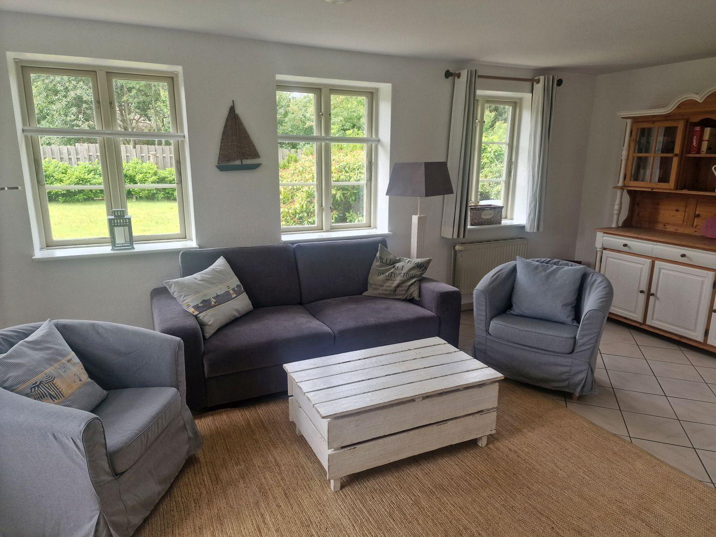 Wohnzimmer mit Sofa, zwei Sesseln und Holztisch. Fenster zeigen Garten. Wanduhr und Schrank sichtbar.
