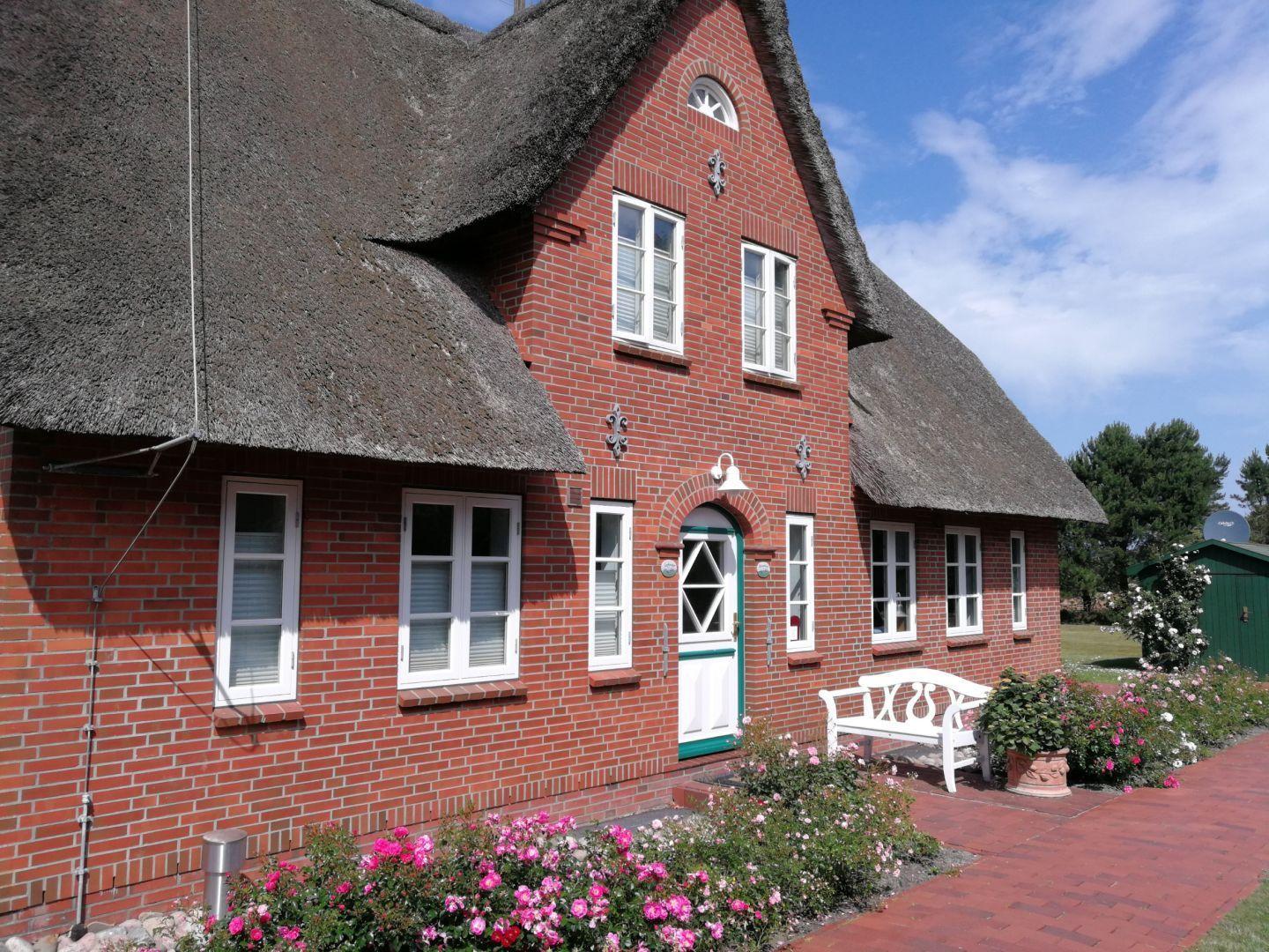 Rotes Backsteingebäude mit Strohdach, weißer Tür und Blumenbeete.