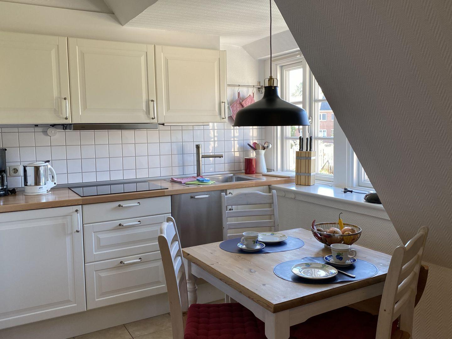 Küche mit Esstisch, weißen Schränken und Fenster. Tisch mit Tassen und Obstschale.