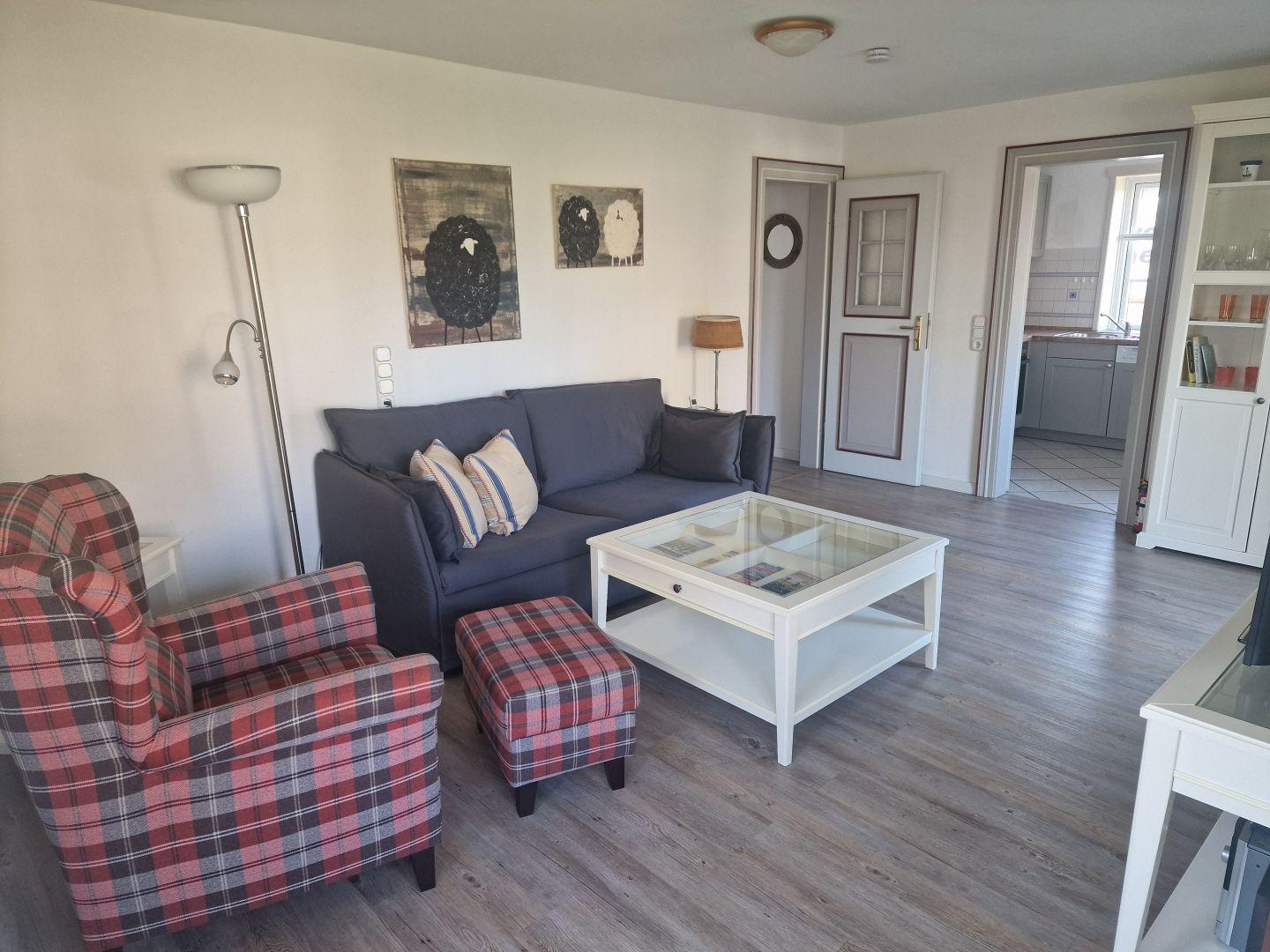 Wohnzimmer mit Sofa, Sessel und Glas-Tisch. Küche und Flur sind sichtbar.