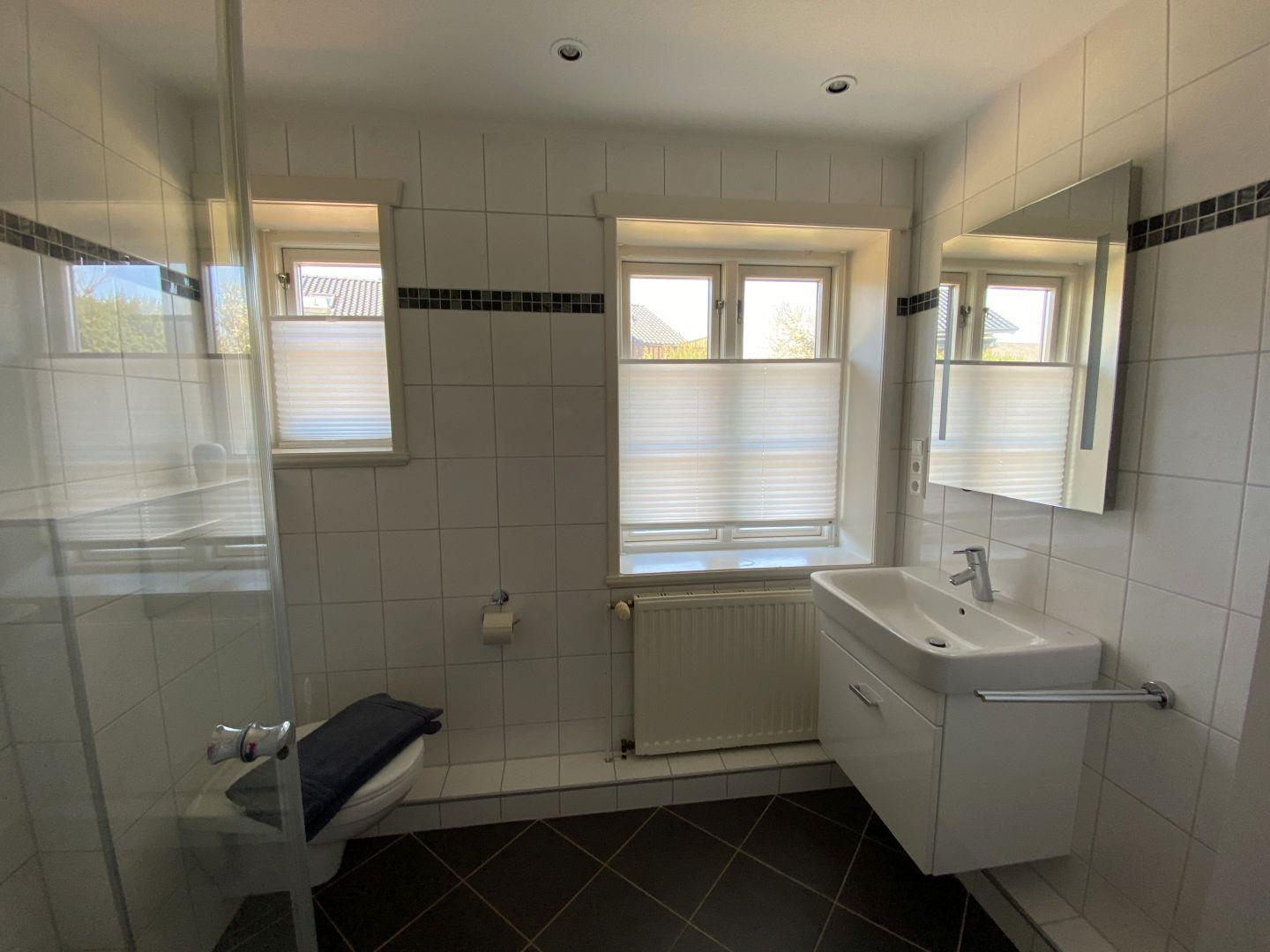 Badezimmer mit Dusche, Waschbecken und Toilette. Fenster mit Jalousien.