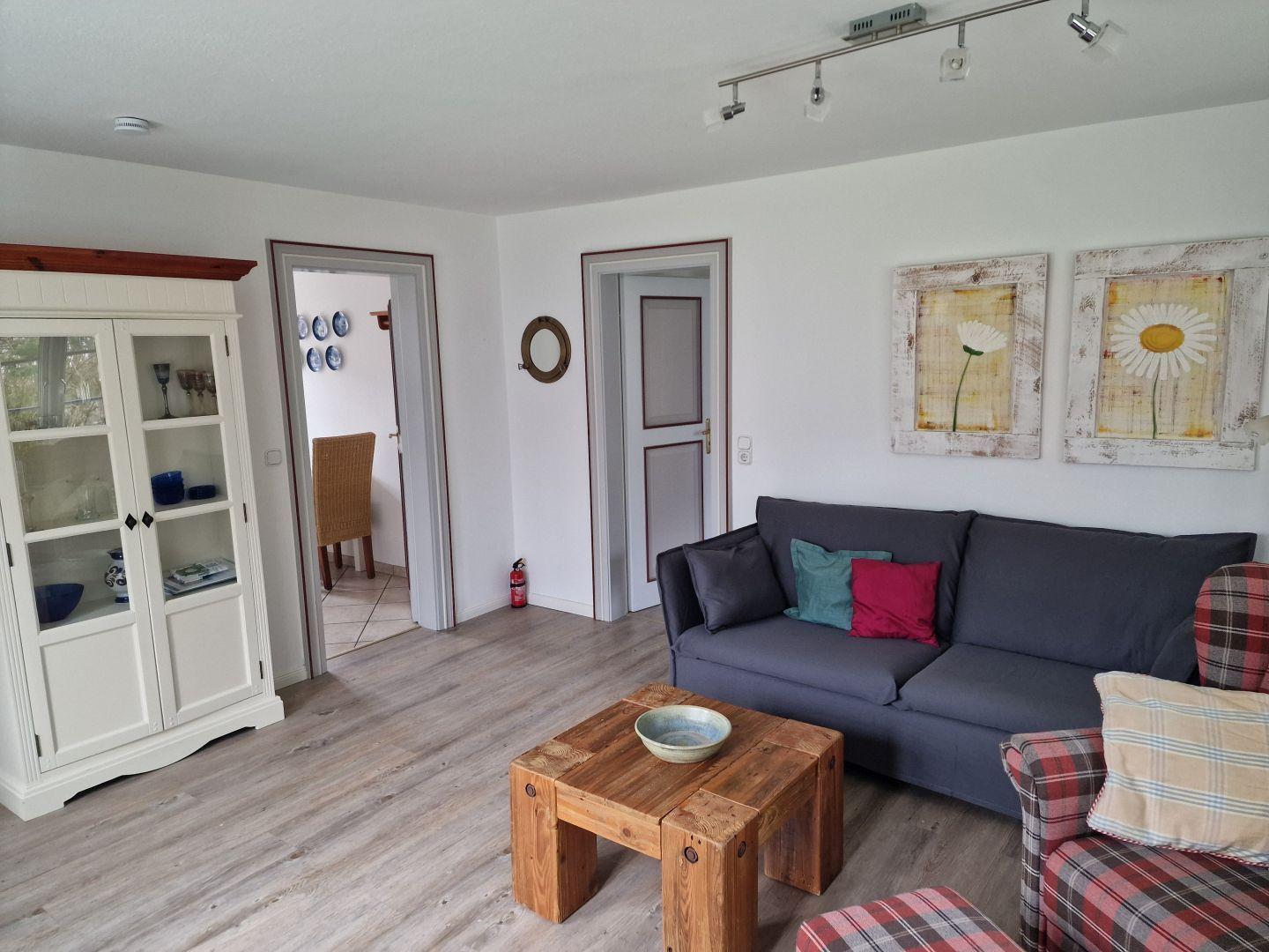 Wohnzimmer mit Sofa, Couchtisch und Schrank. An der Wand hängen zwei Blumenbilder.