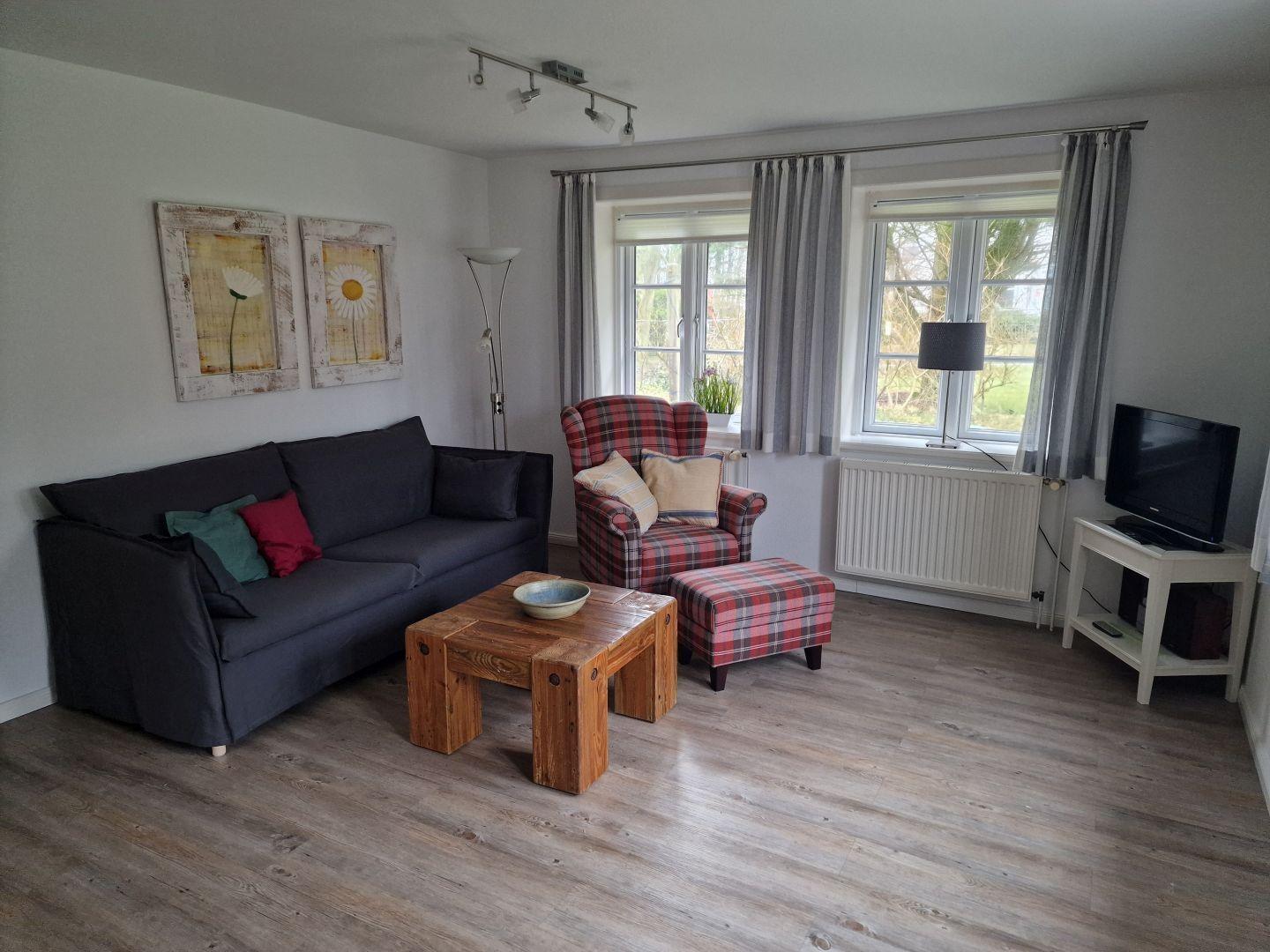 Wohnzimmer mit Sofa, Sessel und Holztisch. Fenster mit Vorhängen und Fernseher.