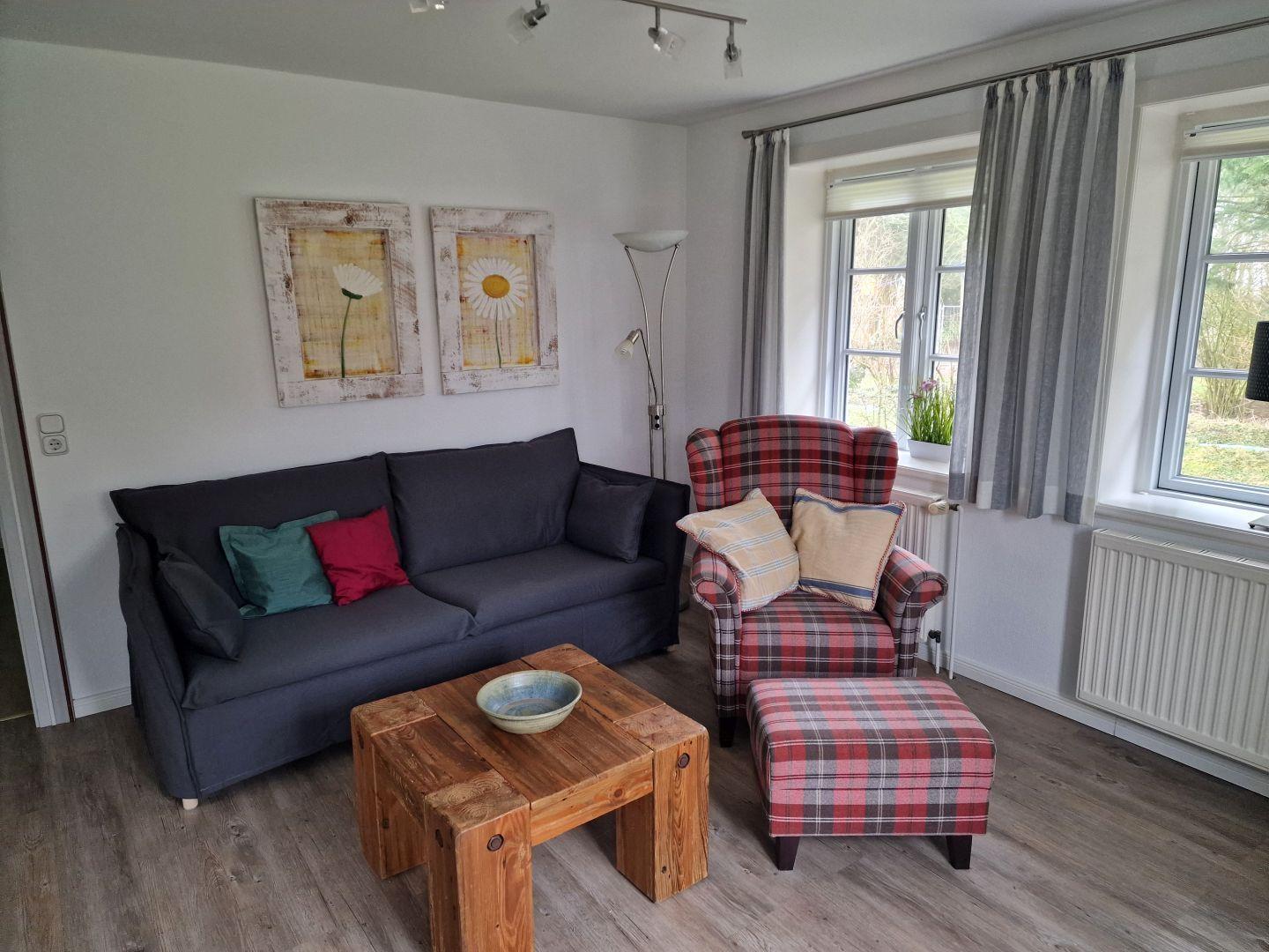 Wohnzimmer mit Sofa, Sessel und Holztisch. Fenster mit Vorhängen und Wandbildern.