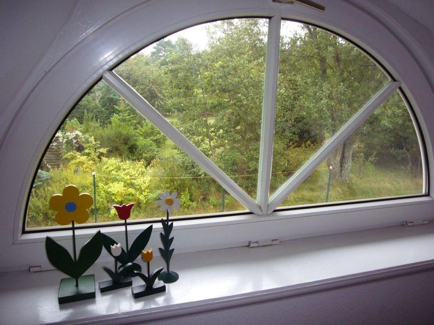 Blick durch ein rundes Fenster auf einen grünen Garten mit Blumenfiguren auf dem Fensterbrett.