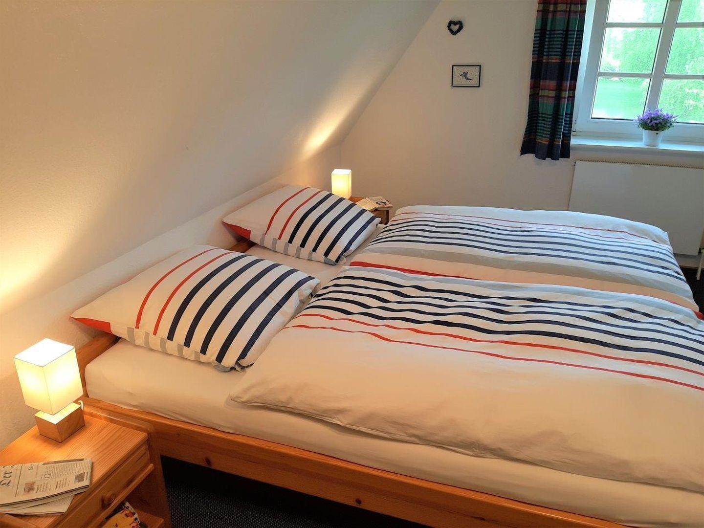 Zimmer mit Doppelbett, Nachttisch und Fenster. Bett mit Streifenbezug.