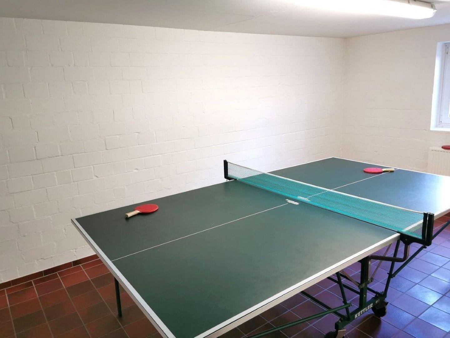 Tischtennisplatte mit Netz und Schlägern in einem Raum mit weißen Wänden und gefliestem Boden.